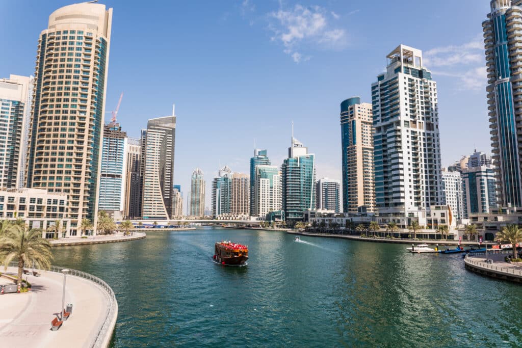 A traditional wooden boat sails through the Dubai Marina canal, surrounded by modern high-rise buildings and palm-lined walkways under a clear blue sky.